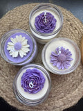 Four small clear glass candles arranged on a woven beige mat. Three candles feature sculpted purple flower tops, and one candle has a white daisy flower top with a purple center.