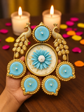A decorative gold-toned Diwali lamp holder with peacock motifs and five light blue circular panels, held in a hand over a wooden surface with lit candles and colorful flower petals in the background.