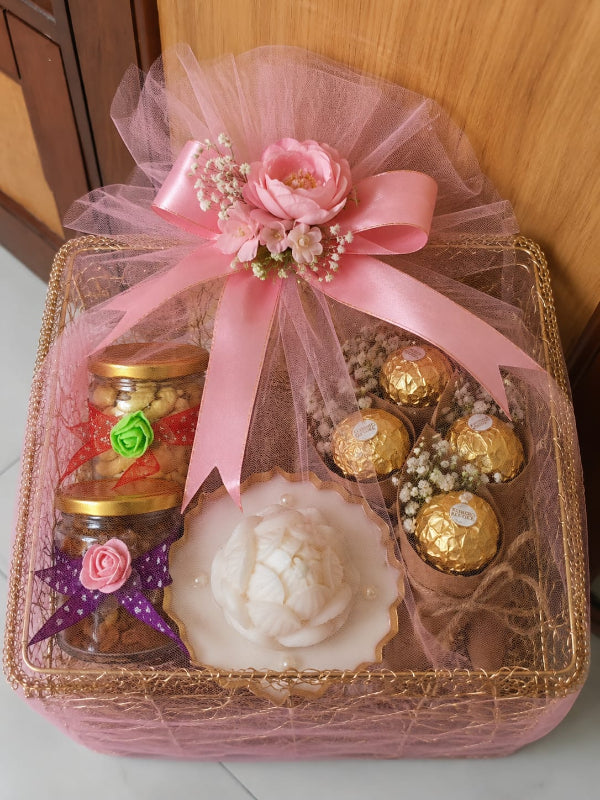 A pink and gold gift basket containing a white rose-shaped candle, two small jars with gold lids, and four Ferrero Rocher chocolates, decorated with a large pink ribbon, artificial flowers, and baby's breath on a pink tulle wrap.