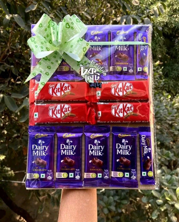 A gift basket filled with assorted chocolate bars, including Cadbury Dairy Milk and KitKat, wrapped in clear plastic and tied with a green patterned bow, held up by a hand against a background of green foliage.