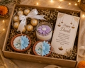 A Diwali gift box filled with festive treats: two decorated blue flower candles in brown cups, a glass jar candle with a purple flower, a clear bowl of Ferrero Rocher chocolates tied with a white ribbon, and a printed Diwali greeting card inside a kraft box.