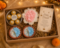 A Diwali gift box set containing Ferrero Rocher chocolates, a pink flower-shaped soap, two blue peacock candles, and a 'Happy Diwali' card, arranged in a brown box with shredded paper on a warm, decorated background.
