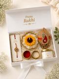 A white gift box tied with a white ribbon, opened to reveal a Rakhi gift set. The set includes a printed Rakhi on a card, a yellow and white flower-shaped diya, a decorative peacock ornament, two red and white candles, and a small jar labeled 'SPECIAL HAPPY Rakhi Day'.