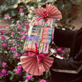 A gift basket filled with assorted chocolate bars, wrapped in pink pleated paper and tied with pink ribbons, held by a person in a garden with pink flowers in the background.
