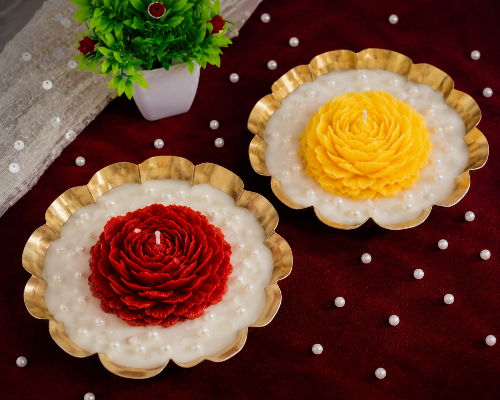 Two decorative candles shaped like flowers on a red surface with a small plant in the background.