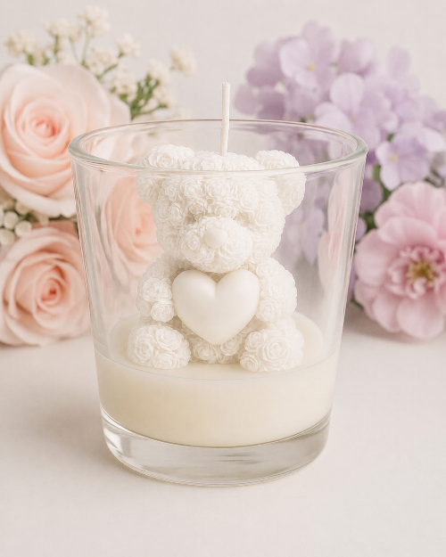 Candle in a glass container with a decorative bear inside, surrounded by flowers on a light background